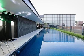 A modern indoor swimming pool with a glass wall, a poolside area with a metal ladder, and a building with large windows in the background. - The Marq Apartment Quan 1  (Ho Chi Minh City)