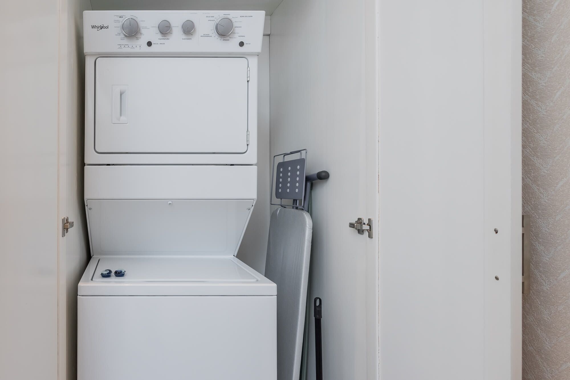 A compact, white laundry room with a stacked washer and dryer, a metal drying rack, and a door with a simple latch.