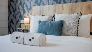 Una cama bien hecha con una toalla blanca enrollada, un almohadón azul y almohadones estampados en una habitación con cabecera de madera y papel tapiz con motivos de hojas.