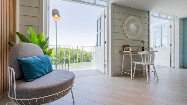 Una sala moderna con un sofá gris, una mesita y una silla. Hay un balcón con vistas a zonas verdes y una masa de agua.