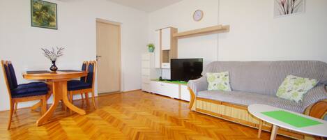 A living room with a wooden herringbone floor, a sofa, a TV, a dining table with chairs, and a small side table.