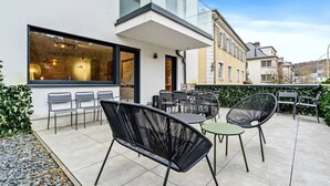 Eine Terrasse im Freien mit moderner Möblierung, einem Glasgeländer und Blick ins Innere eines Gebäudes.