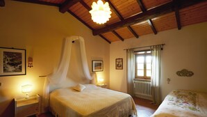 Een slaapkamer met een groot bed, een kleiner bed, een nachtkastje met een lamp, een raam met gordijnen en een houten plafond.