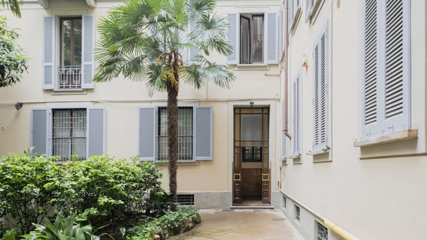 Une petite cour intérieure avec un palmier, une porte dotée d’un panneau de verre, et un bâtiment avec des volets et des balcons.