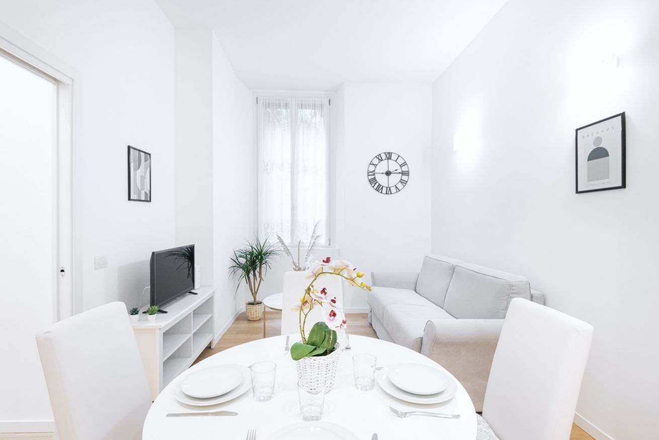 A modern, well-lit living room with a round dining table set for two, a television on a stand, a sofa, and a clock on the wall.