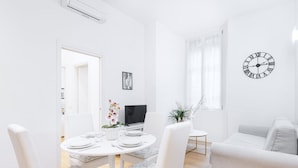 Un salon moderne aux tons clairs, avec un espace repas, un canapé, un téléviseur et une horloge murale.