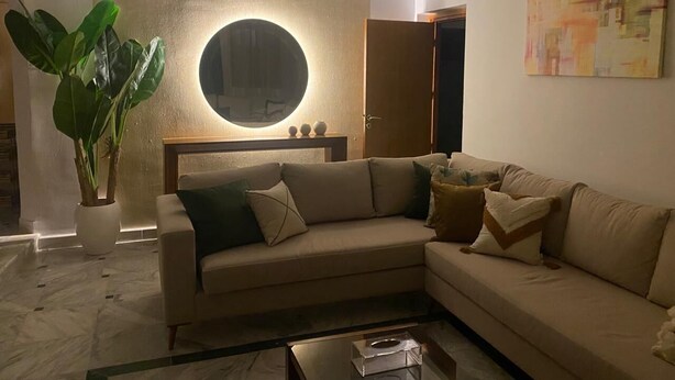 Una sala de estar moderna con un sofá color beige, un espejo redondo, una planta en maceta y un diseño minimalista.