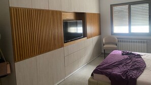 Un dormitorio con una televisión montada en una pared de paneles de madera, una cama con una colcha morada y una silla cerca de una ventana con persianas.