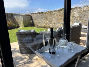 Un patio con muebles de mimbre, una mesa con una botella de vino, copas y vistas a un muro de piedra y jardín.