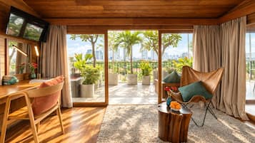 Una sala de estar moderna con techo de madera, televisor, escritorio, silla, sofá y vista de un paisaje urbano a través de grandes puertas de vidrio.