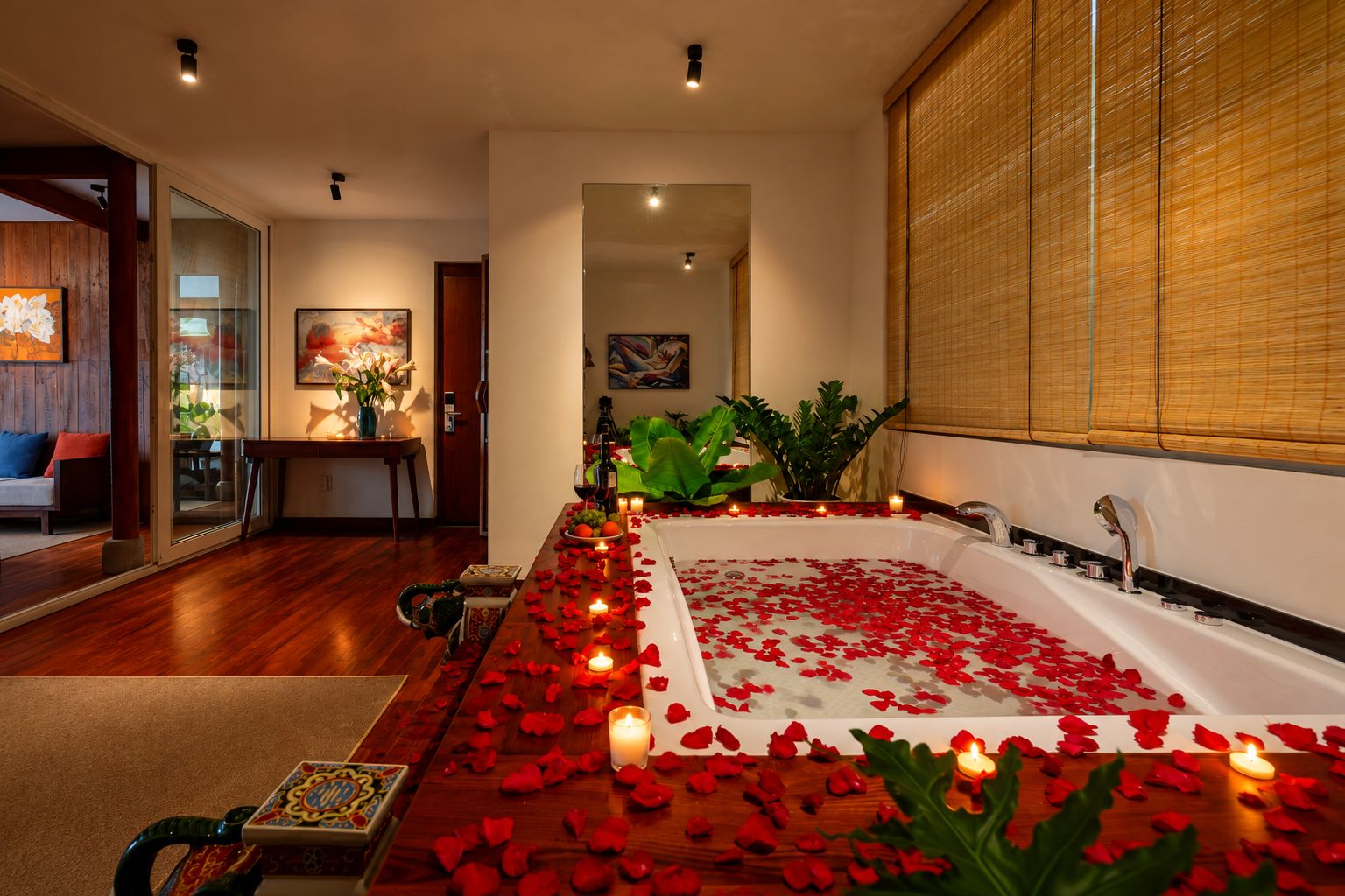 A room with a large bathtub filled with rose petals and candles, a wooden floor, and a view of a cityscape through large windows.