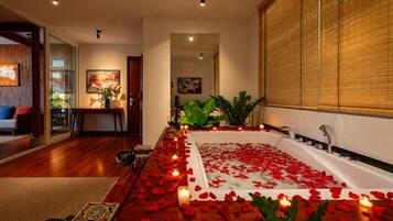 A room with a large bathtub filled with rose petals and candles, a wooden floor, and a view of a cityscape through large windows.