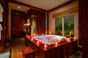 Habitación con una bañera de madera grande rodeada de velas y pétalos de rosa, una mesita con frutas y copas de vino, y vista a la vegetación a través de una ventana.