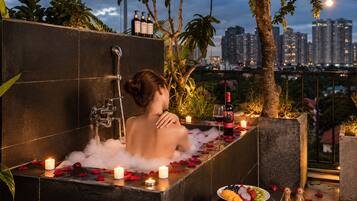Una mujer en una bañera con pétalos de rosa y velas, rodeada de plantas y vista a la ciudad.