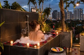 A woman in a bathtub with rose petals and candles, surrounded by plants and a cityscape view. - Nexus House Lang Bao Chi (Ho Chi Minh City)