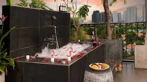 Una bañera negra con pétalos de rosa y velas, rodeada de follaje y vistas a la ciudad.