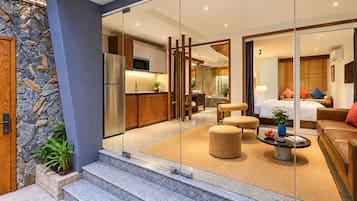 Quarto de hotel moderno com divisória de vidro, cozinha, sala de estar com sofá e banquinho, e banheiro com chuveiro e vaso sanitário.