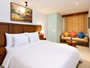 A hotel room with a large bed, a sofa with colorful cushions, a painting on the wall, and a window with blinds. - Nexus House Lang Bao Chi (Ho Chi Minh City)