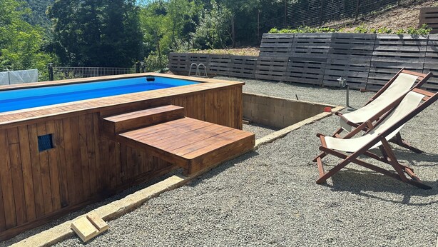 Uma piscina de madeira com escadas para acesso, rodeada por cascalho. Há espreguiçadeiras e um deck de madeira.