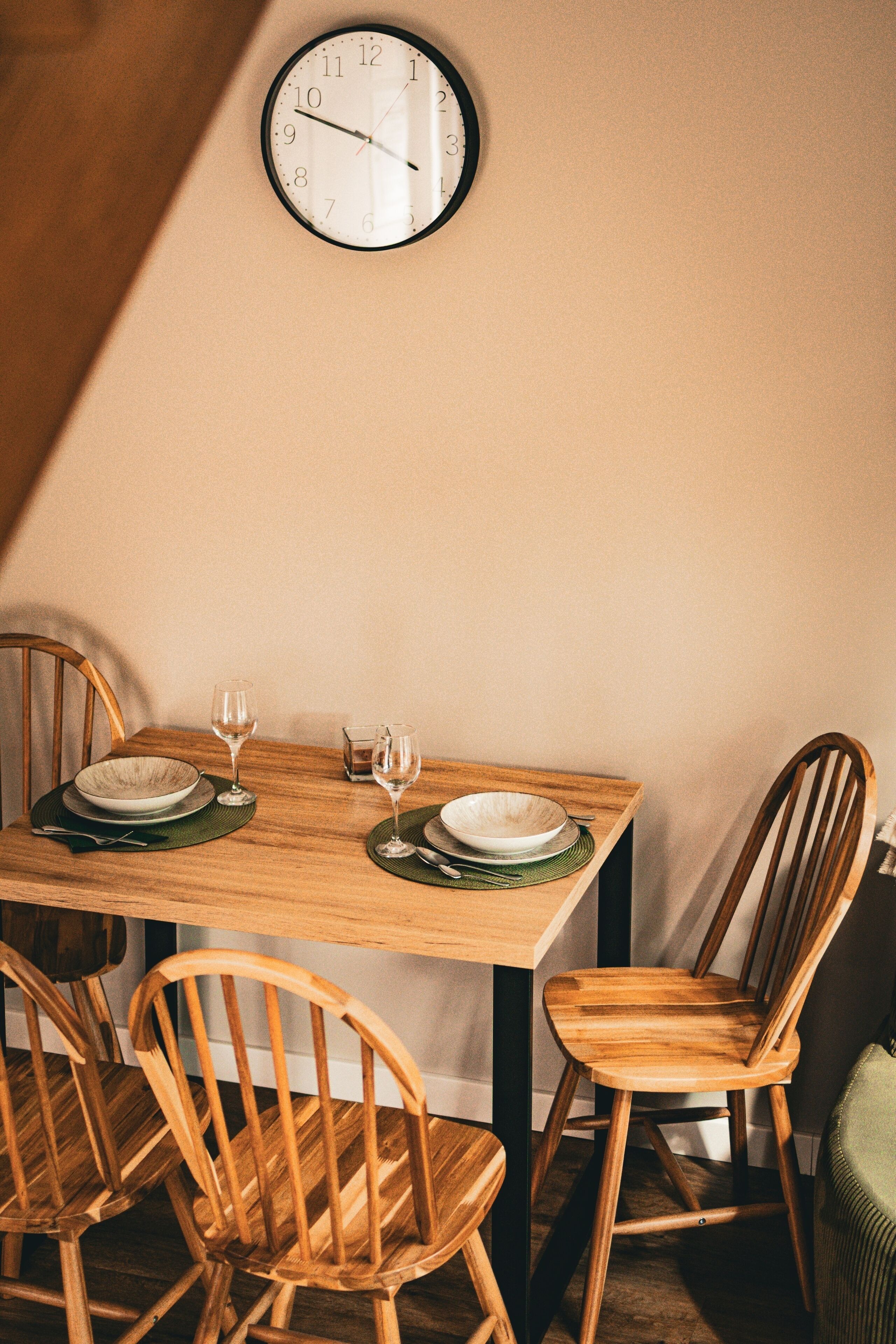 A dining table with four wooden chairs, a clock on the wall, and two wine glasses on the table.
