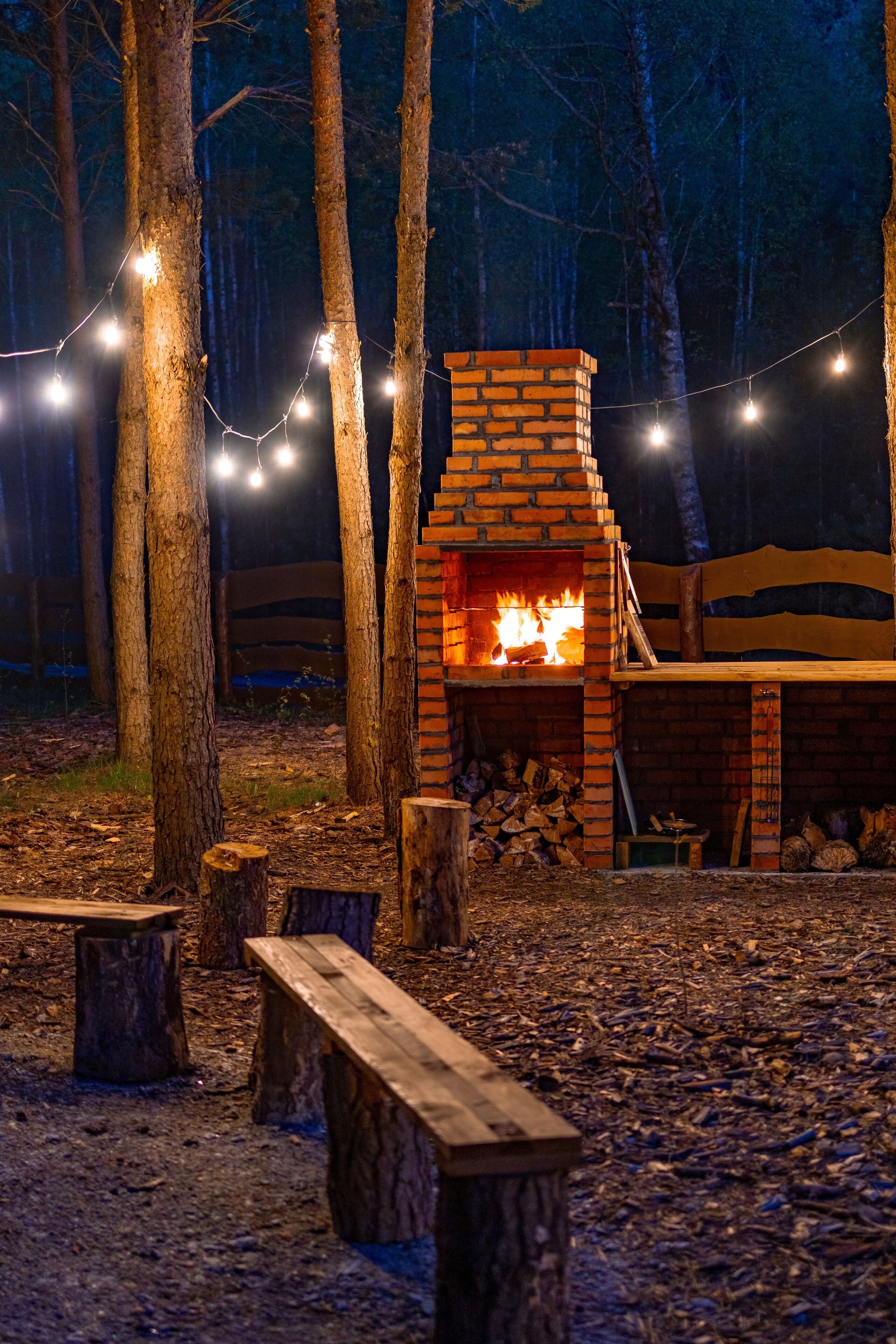 A rustic outdoor fireplace surrounded by trees, with string lights above.