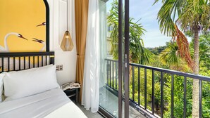 Ein Hotelzimmer mit Bett, Nachttisch, Fenster mit Blick auf Palmen und Grünpflanzen sowie ein Balkon mit Geländer.