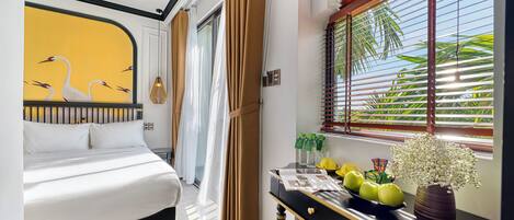 A hotel room with a bed, a desk with a vase and fruits, and a window with blinds.