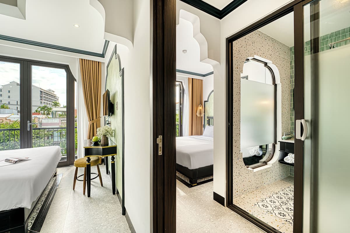 A hotel room with a bed, a desk, a chair, a bathroom with a shower, and a balcony with a view of buildings.