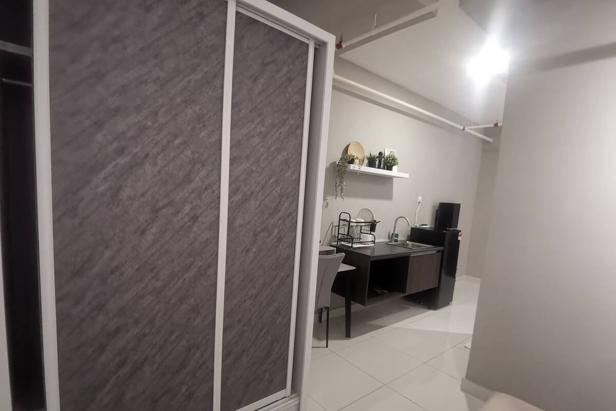 Cozinha moderna com bancada escura, armários brancos e geladeira preta. Há uma pia branca com torneira e uma pequena prateleira acima, com objetos decorativos.