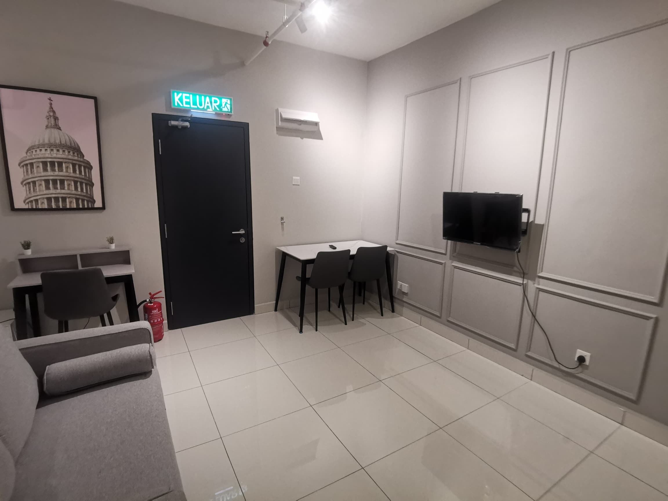 A modern living room with a black door marked "KELUAR," a dining area with a table and chairs, a sofa, a TV mounted on the wall, and a framed picture of a building.