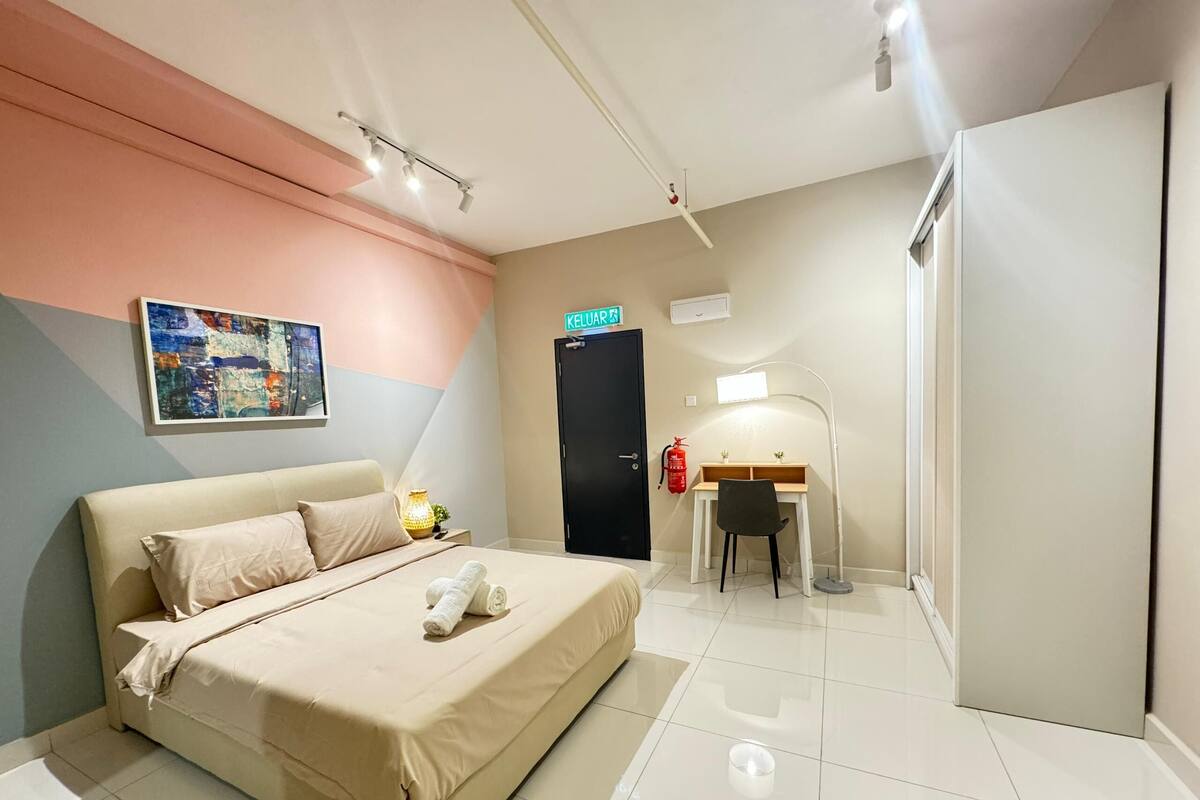 Quarto de hotel com cama, escrivaninha, cadeira, abajur, extintor de incêndio e um quadro na parede.
