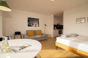 Una habitación de hotel moderna con una cama, una mesa de comedor redonda, un sofá con cojines amarillos, un cuadro en la pared y una kitchenette al fondo.