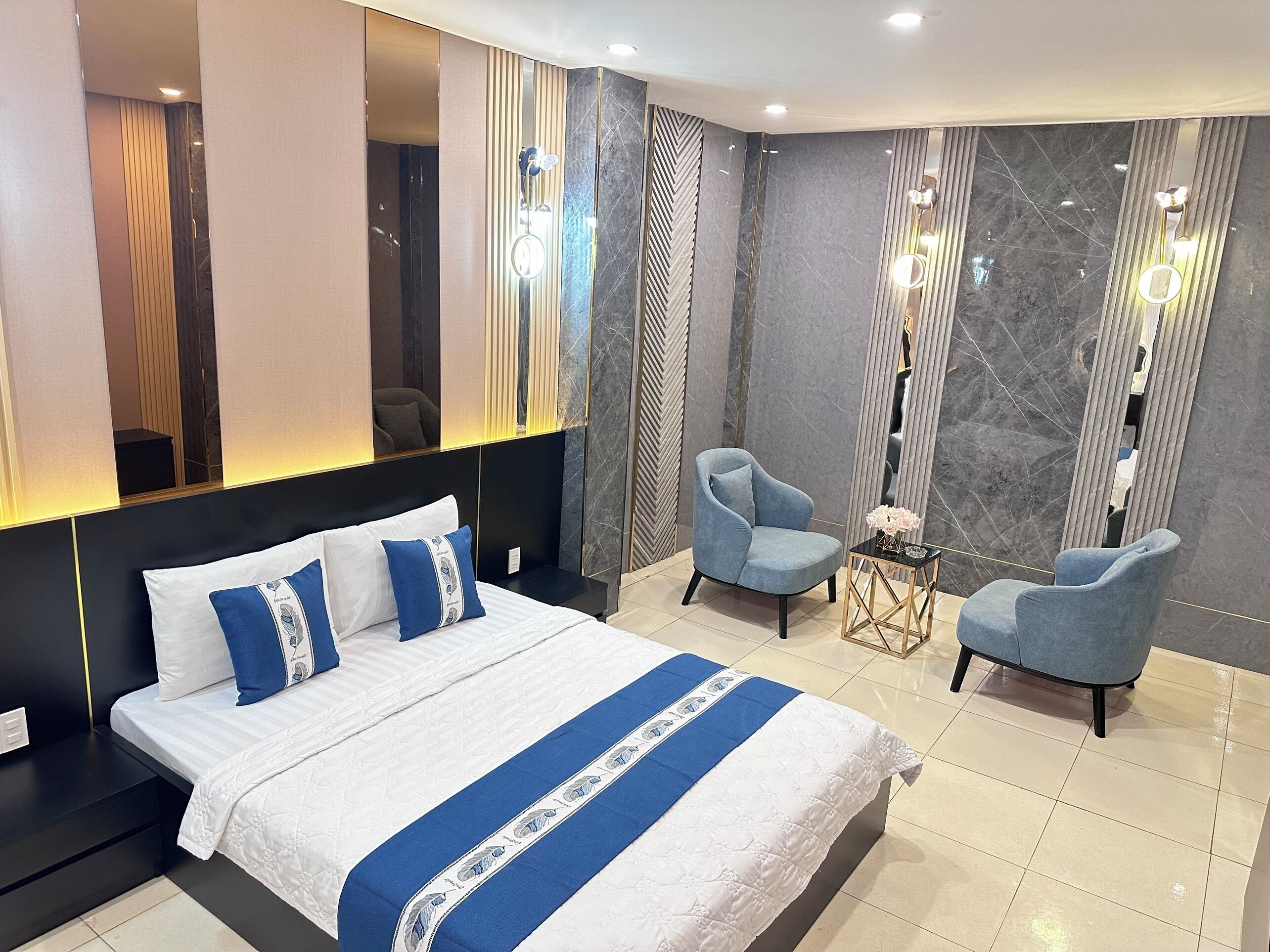 A modern hotel room with a large bed, two armchairs, a small table, and a mirror.