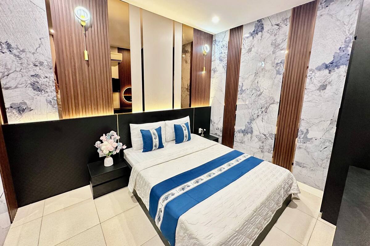 A modern bedroom with a large bed, a nightstand, a vase of flowers, and a wall with vertical wooden panels and a circular light fixture.