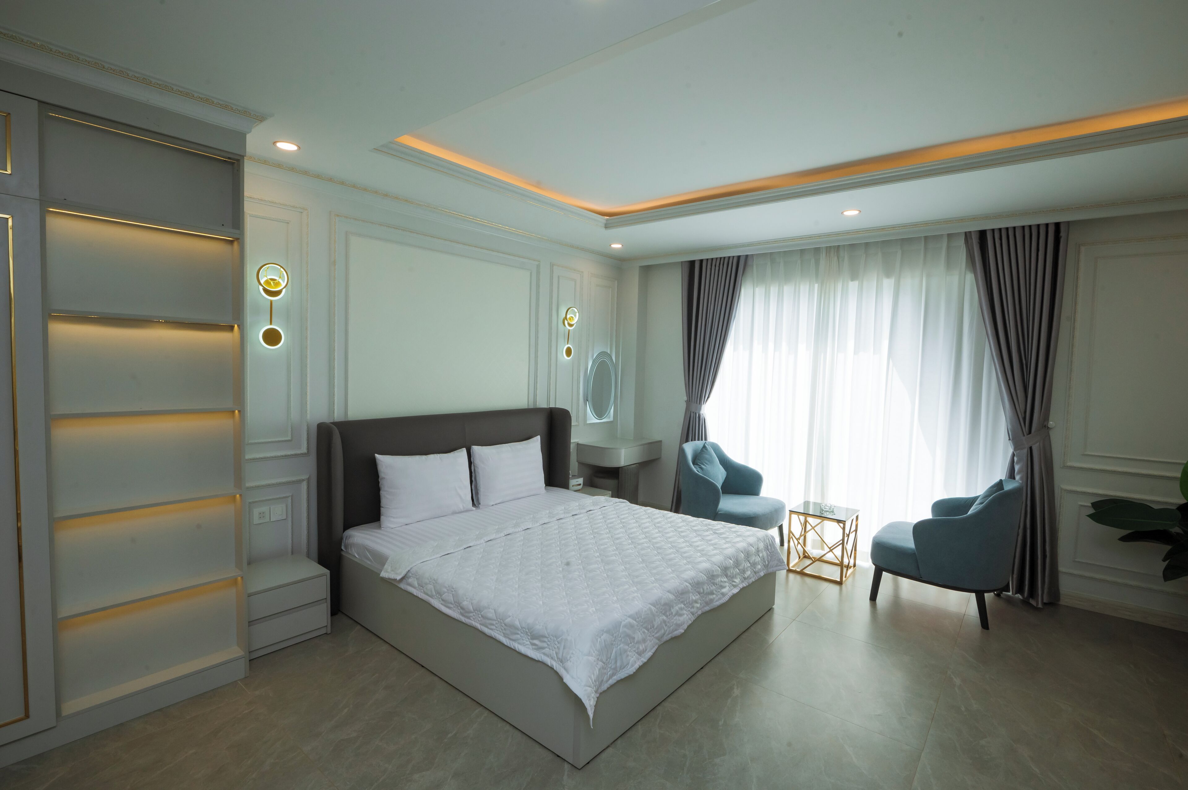 Un dormitorio de hotel moderno con una cama grande, una zona de estar con dos sillones, una mesita y un ventanal con cortinas.