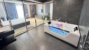 Une salle de bain moderne dotée d’une grande baignoire, d’une douche séparée par des parois en verre et d’une chambre à coucher visible, avec un lit et des rideaux.