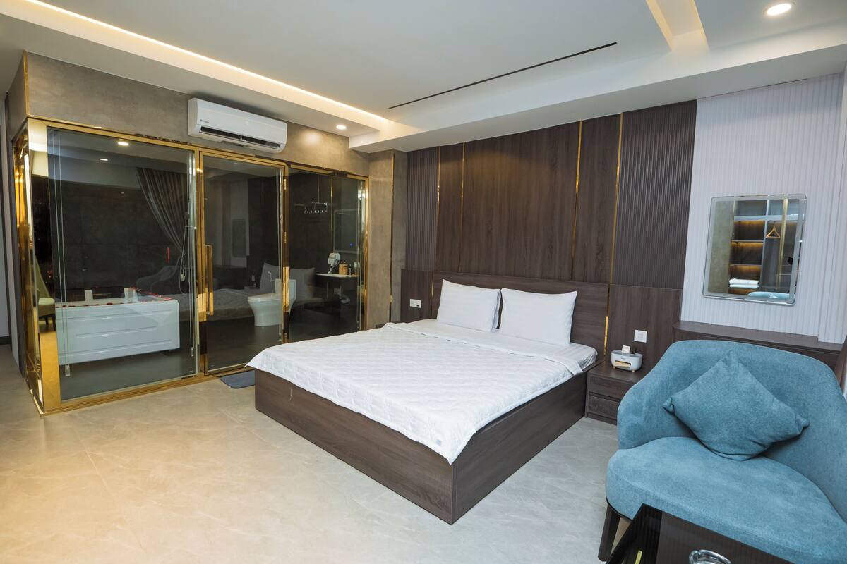 A modern bedroom with a large bed, a glass-enclosed walk-in closet, a seating area, and a built-in wardrobe.