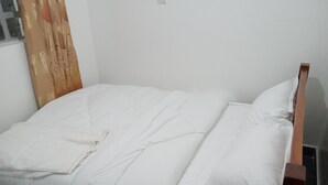 Una cama individual con ropa de cama blanca en una habitación con una pared blanca lisa y una ventana con cortina.