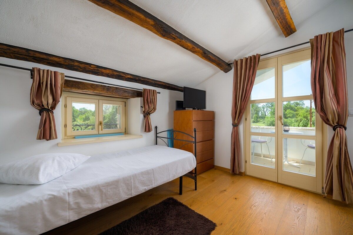 A room with a bed, a television on a wooden cabinet, a window with curtains, and a sliding glass door to an outdoor area.