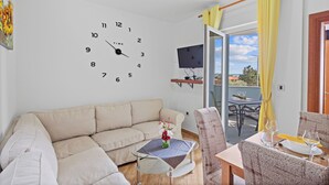 Ein Wohnzimmer mit einem beigen Sofa, einem Glastisch mit Blumenvase, einem Esstisch mit Stühlen und einer großen Wanduhr.