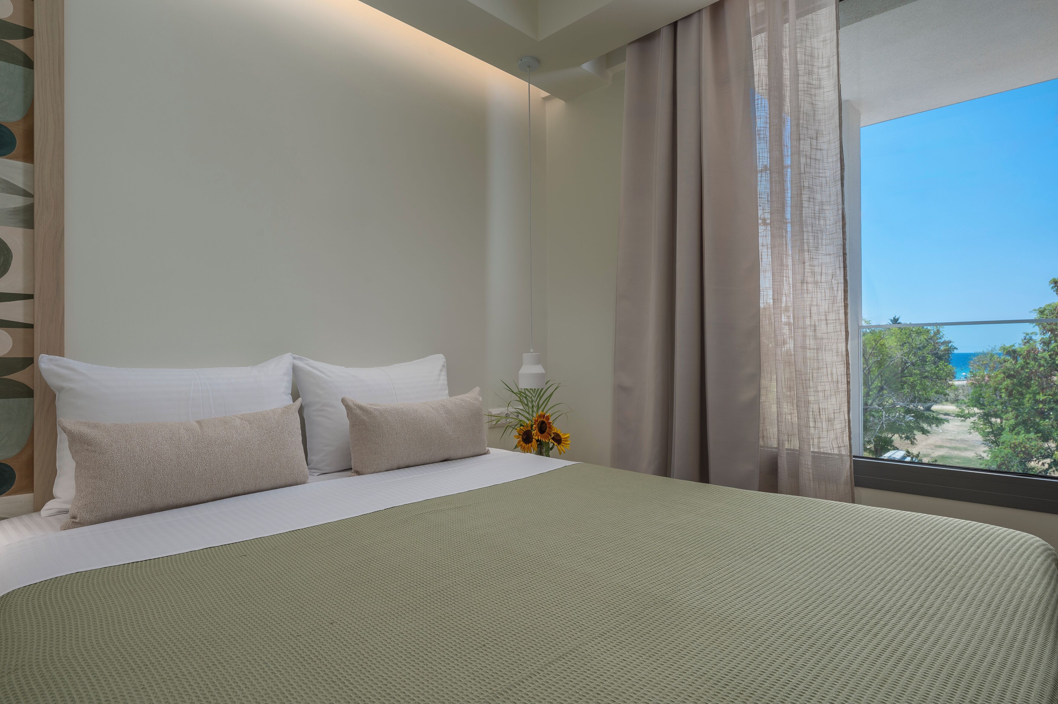 A hotel room with a large bed, a nightstand with a lamp, and a window with a view of trees and a blue sky.
