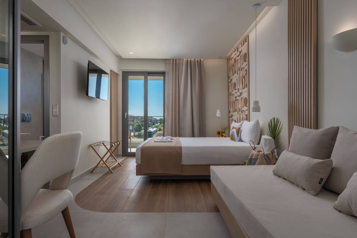 A modern hotel room with a bed, a sofa, a TV, and a balcony with a city view.