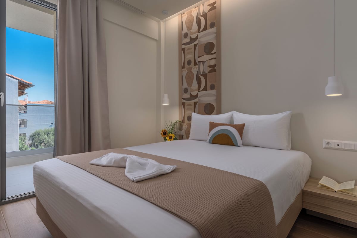 A hotel room with a large bed, a view of the sea, a wall with abstract art, a nightstand with a book, and a window with curtains.