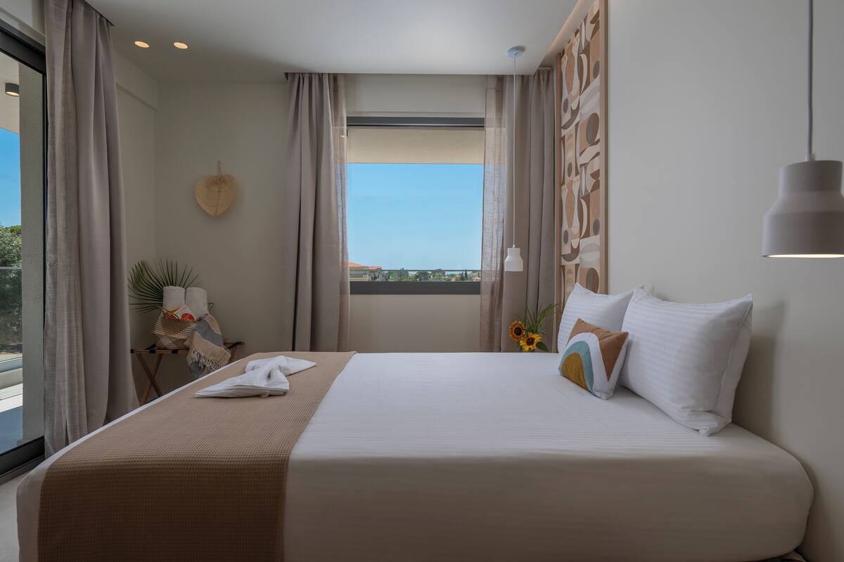 A neatly made bed with a white and beige bedspread, a small table with a towel and a plant, and a window with a view of buildings and trees.