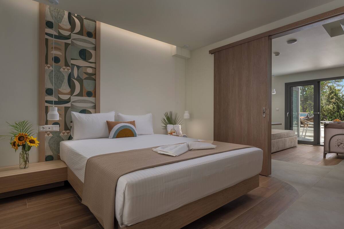 A modern hotel room with a large bed, a wooden wardrobe, a bedside table with a lamp, and a view of the outdoors.
