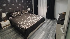 A bedroom with a bed, a nightstand, a dresser, a window with curtains, and a wall with a patterned wallpaper. - Lovely Urban Retreat (București)