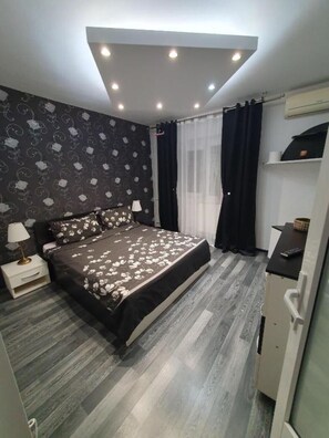 A bedroom with a bed, a nightstand, a dresser, a window with curtains, and a wall with a patterned wallpaper. - Lovely Urban Retreat (București)