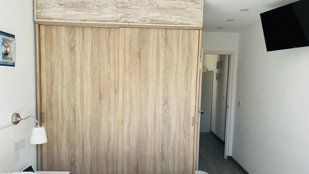 Een hotelkamer met een houten garderobe, een bed met wit beddengoed, een bedlampje en een tv aan de muur.