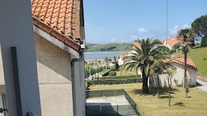 Uitzicht vanaf een balkon op een verhard gebied met een betegeld dak, een waterpartij en een heuvelachtig landschap met palmbomen.