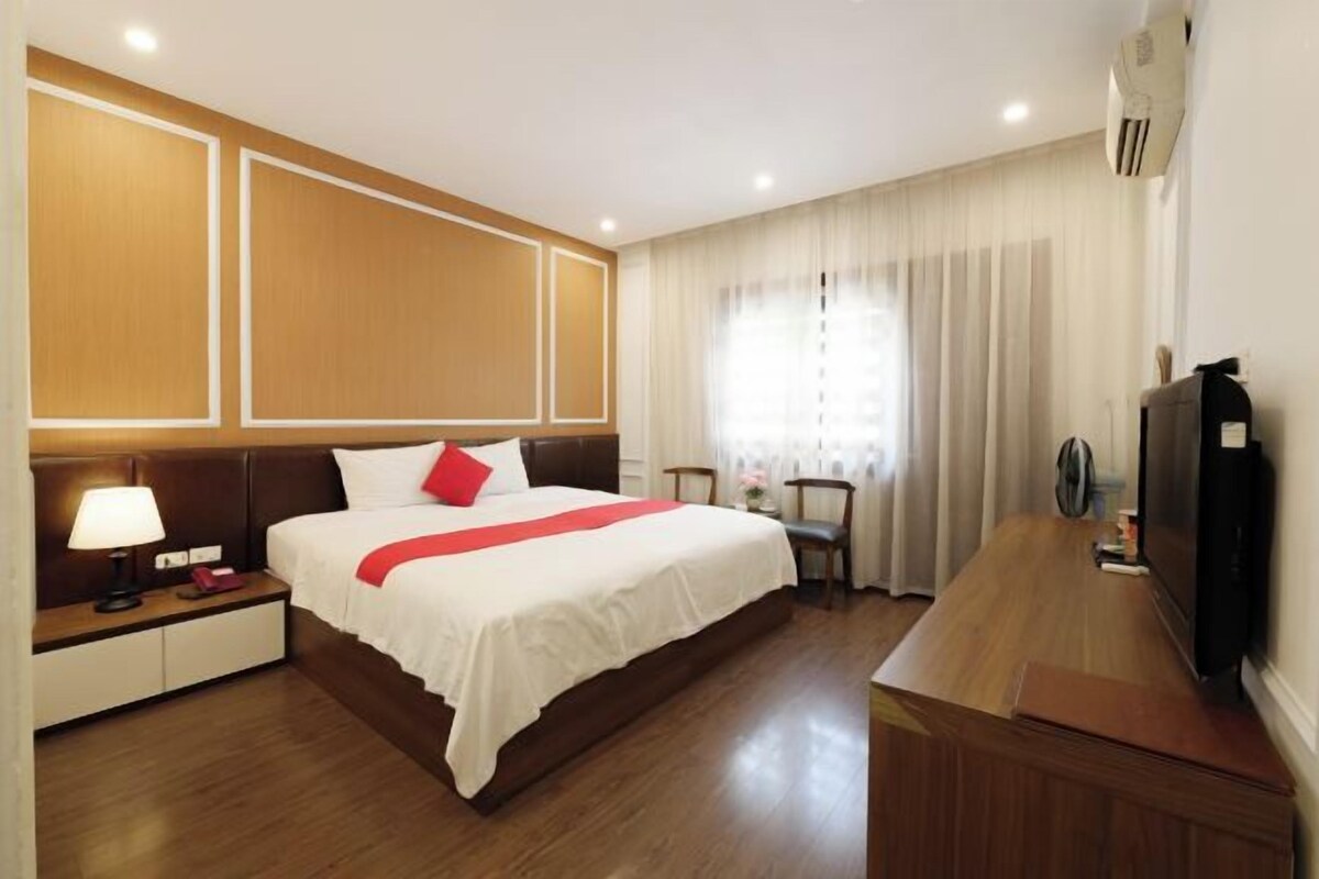 A hotel room with a bed, bedside table, lamp, TV, and a wooden desk.